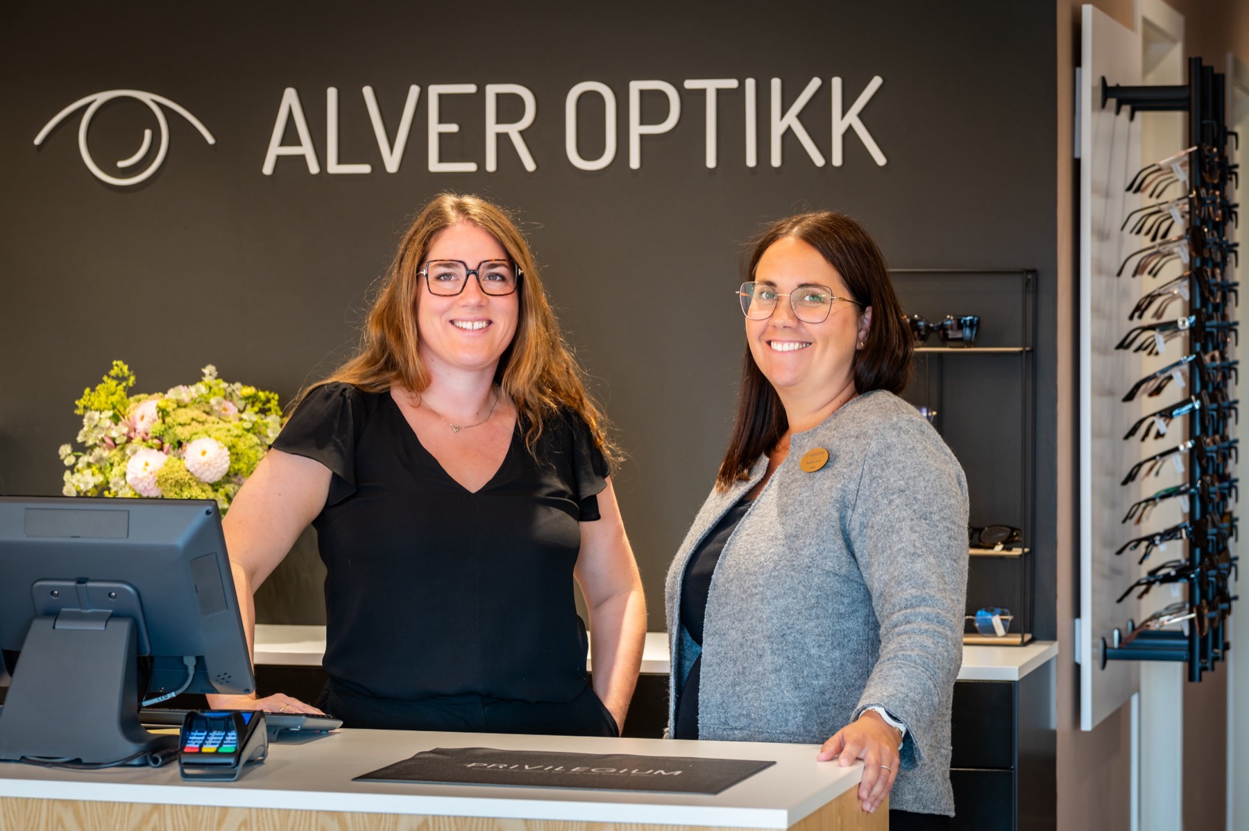 Dei to optikarane i Alver Optikk står bak disken under logoen på veggen, med brillehyller til høgre